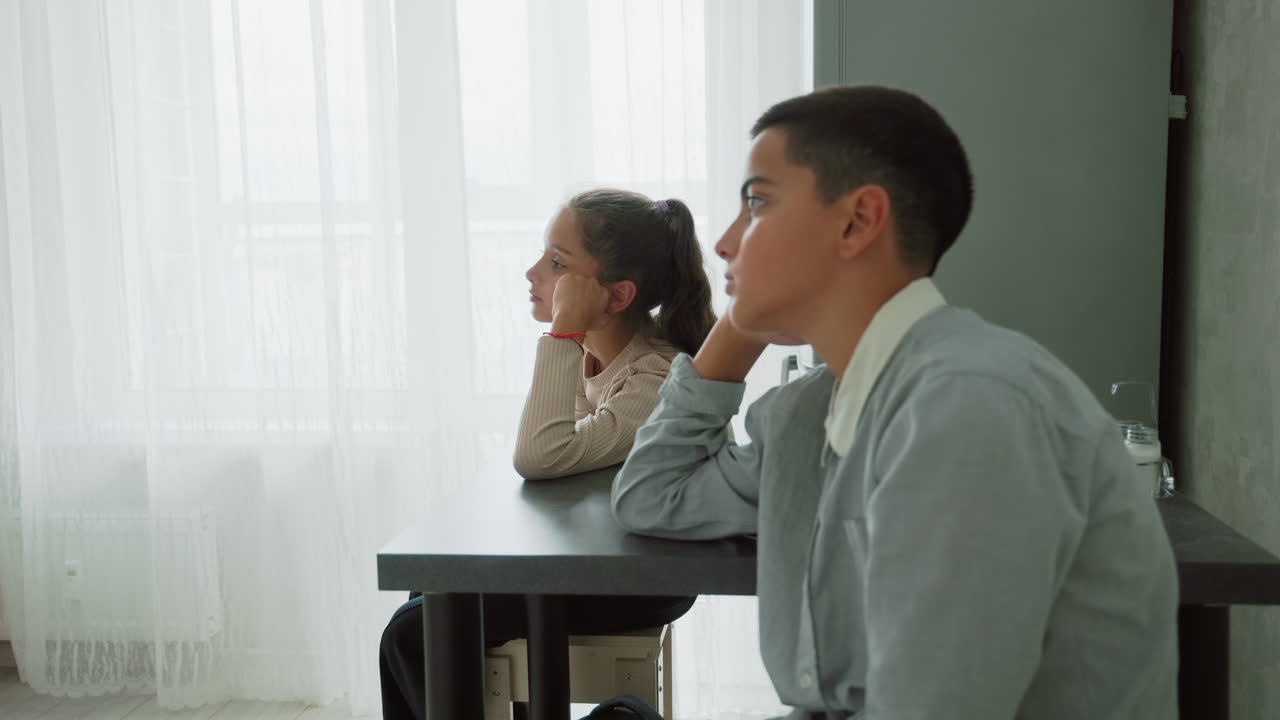 boy and girl in school uniforms sitting quietly at table near sheer curtain window bathed in soft daylight with thoughtful expressions waiting indoors during calm morning home routine moment