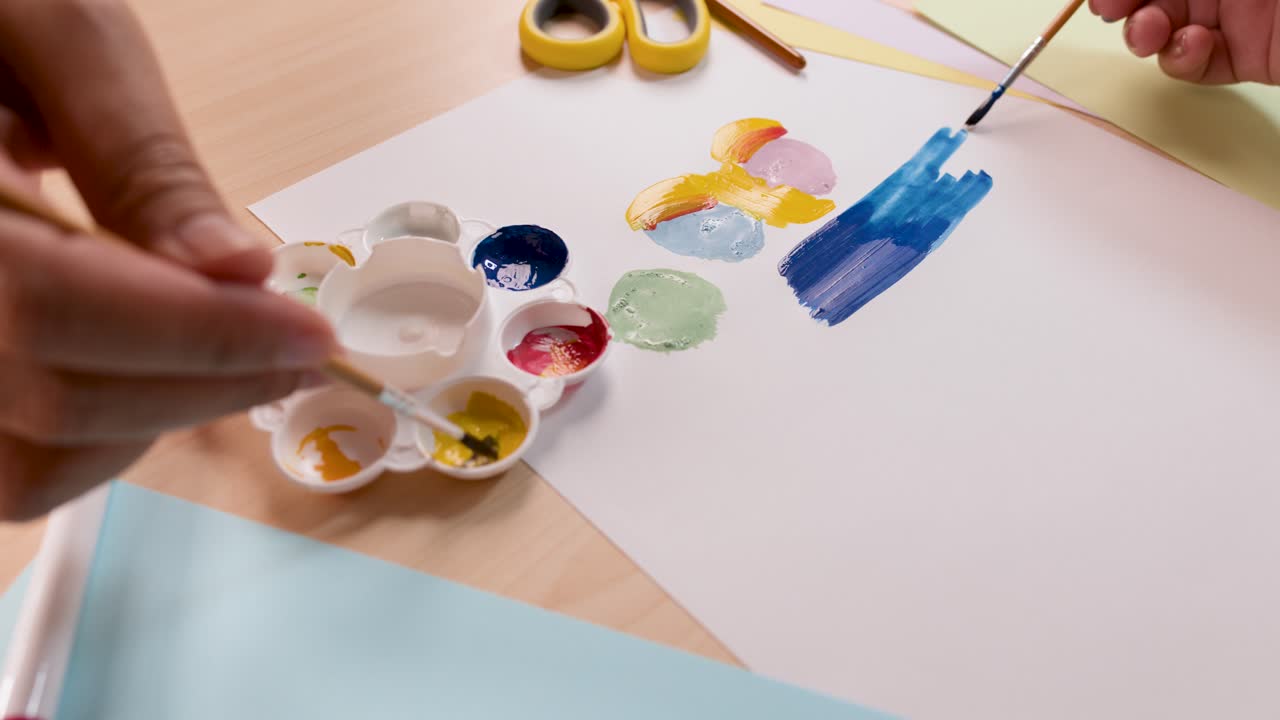 Child’s hands painting with brushes and watercolors on white paper in bright, natural lighting