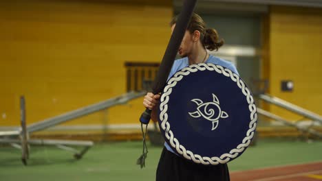 Young-man-with-long-hair-demonstrates-LARP-foam-combat-holding-a-shield-and-a-sword-inside-a-gym