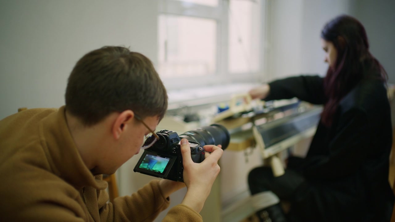 Bak kulissene-opptak av en fotograf som holder et kamera mens han filmer en kvinne som arbeider ved en flatstrikkemaskin nær et lyst vindu.