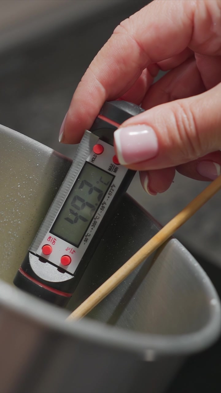 Primo piano di una mano che tiene un termometro digitale in un liquido caldo che mostra cambiamenti di temperatura da 45,1 C a 53,6 C. La scena include goccioline d'acqua visibili e parte di un contenitore di metallo.