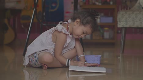 A-young-Asian-girl-sitting-on-the-floor-drawing-in-her-coloring-in-book-with-a-crayon-while-cheerfully-talking-to-herself