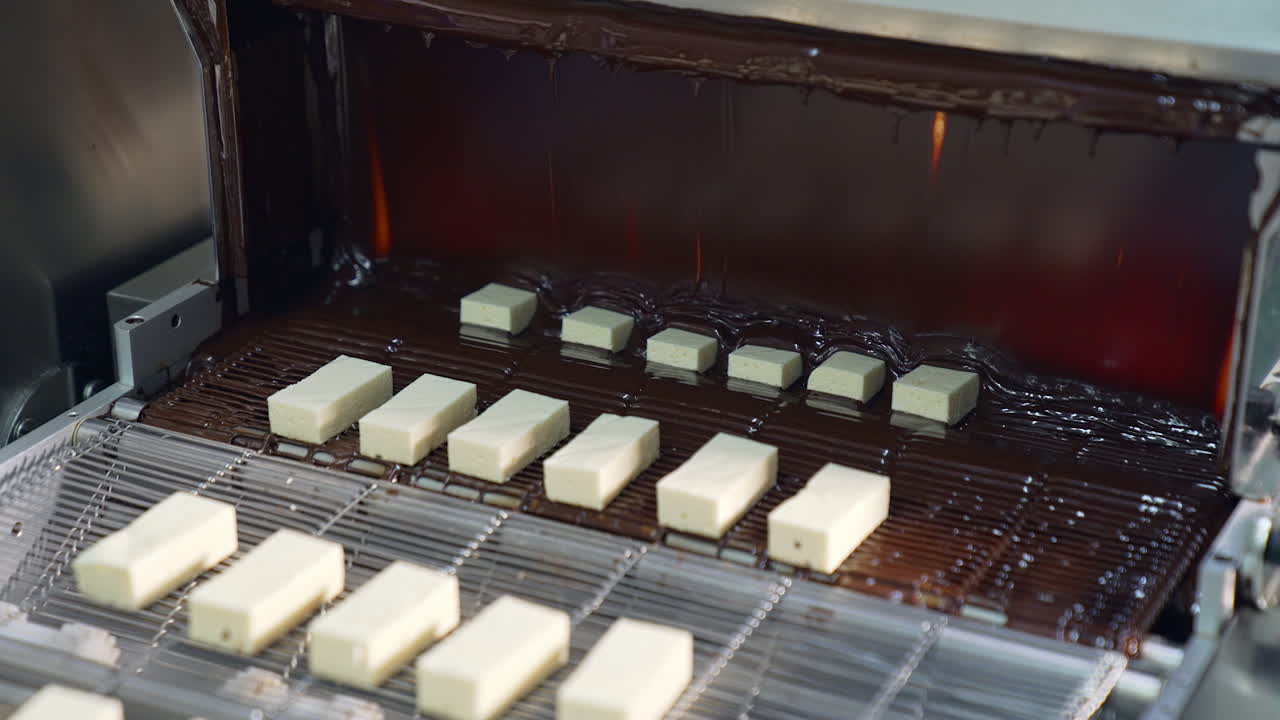 White pieces of desserts being covered with black chocolate. Conveyor belt with sweets goes under the flow of chocolate. Pigeon's milk candies production close up.