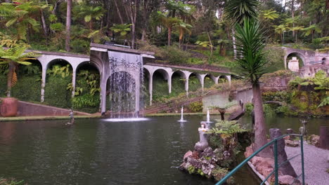 Dolly-shot-of-flowing-waterfall-into-natural-pond-surrounded-by-tropical-plants-and-trees-in-botanic-garden