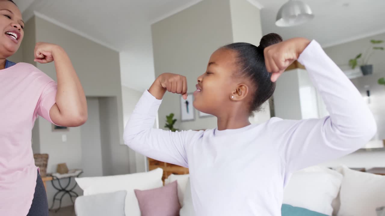 Happy unaltered african american mother and daughter flexing muscles in living room, in slow motion