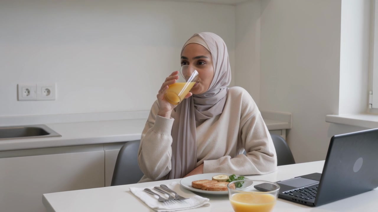 Bringing glass, woman hijab top sipping orange juice at kitchen glancing at laptop, toast for break
