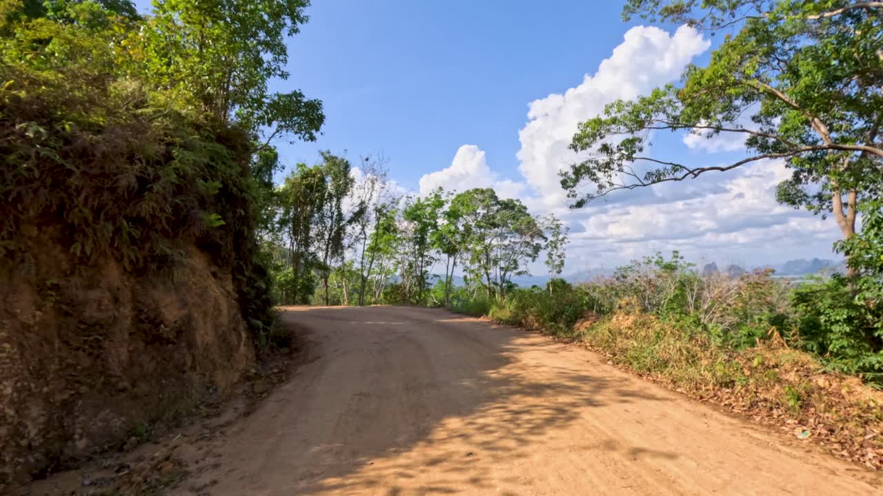 Vehicle travels on winding dirt road through lush, sunlit tropical forest with mountain views