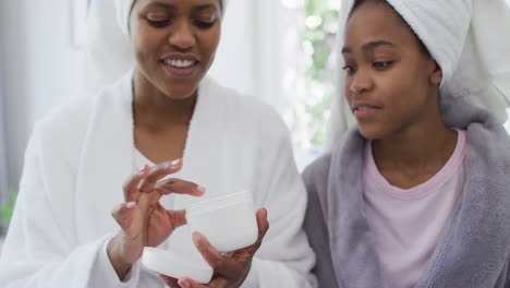 Happy-african-american-mother-and-daughter-wearing-towels-on-head-applying-cream-on-face