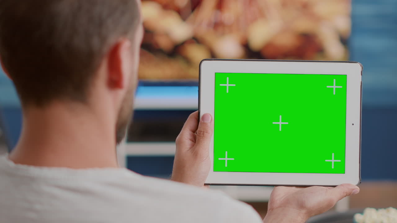 Closeup of man holding digital tablet with green screen watching webinar sitting on sofa