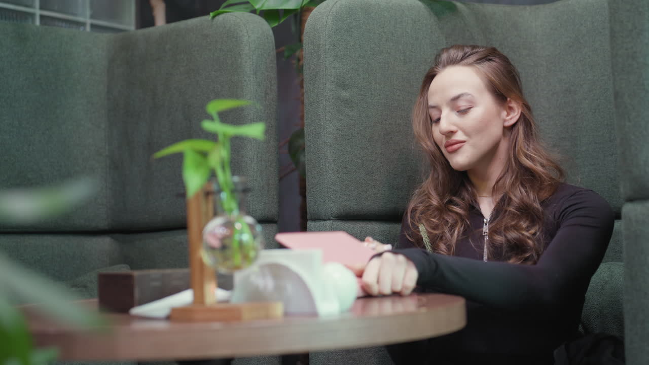 Blonde woman dressed in black outfit sits comfortably in green booth holding pen and preparing to write, surrounded by soft indoor lighting, tabletop decoration, and lush green plants