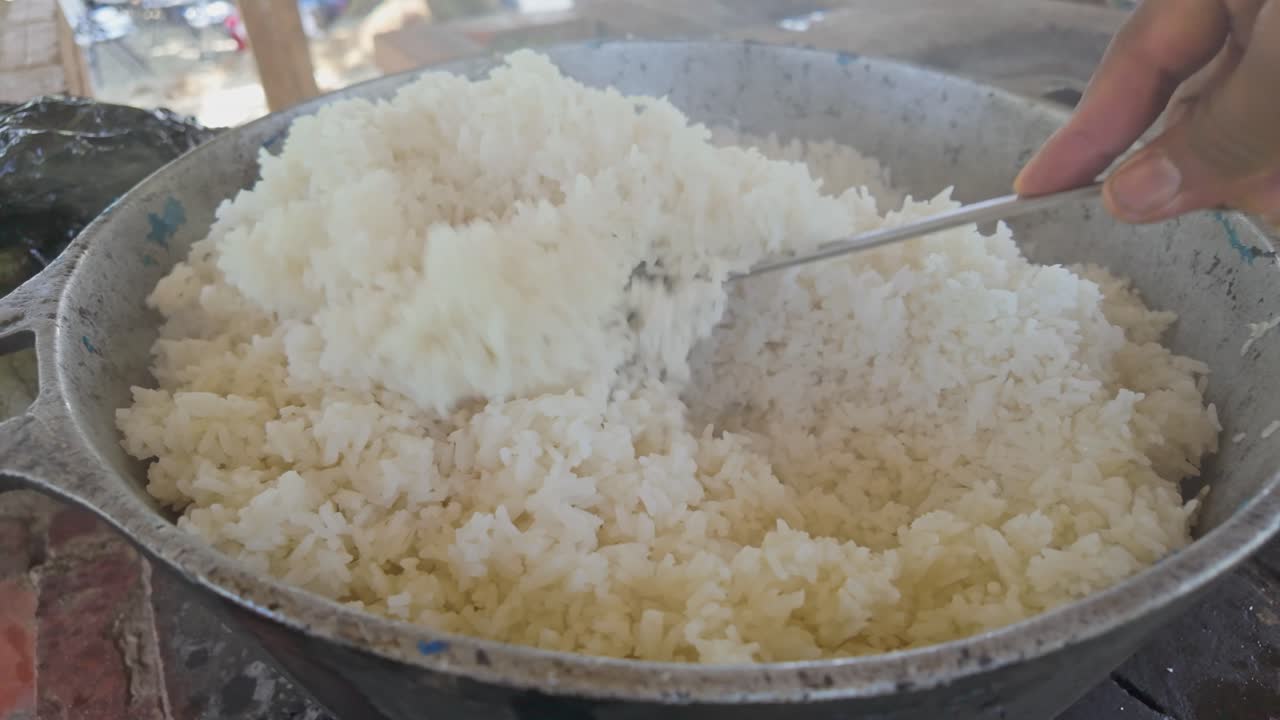 arroz cocinado con leña en un recipiente de hierro, agitado con una cuchara grande, emitiendo vapor en un día soleado en el campo