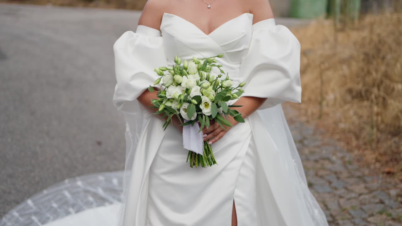 Bride holding a white and green bouquet, wearing an off shoulder wedding gown with a train, standing outdoors on a cobblestone path