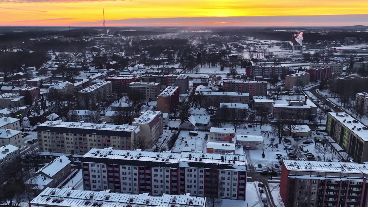 Sneklædt by med beboelsesejendomme og rygende skorstene ved en gylden solnedgang.