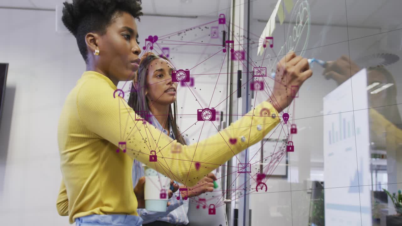 Two women selecting marker and sketching network design on board, pointing laptop as icons floating