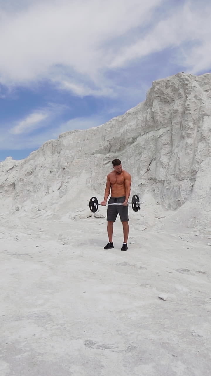 Young athlete lifting heavy barbell on the natural mountain background. Concentrated man engaged with barbell outdoors in a summer day. Vertical video