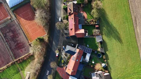 aerial flyover single family houses with solar panels on roof be tennis field during sunny day. fall season in america near rural countryside. car on street in american neighborhood community.