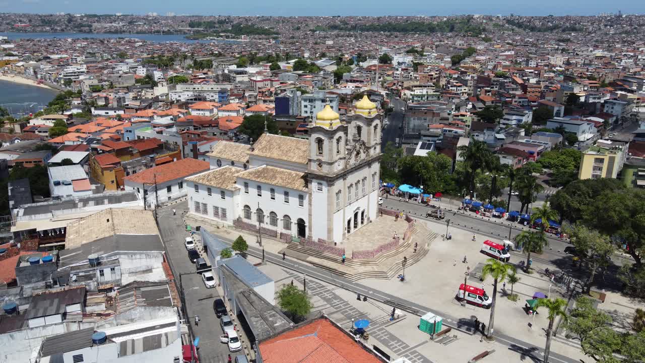 A smooth aerial drone shot circling the historic Bonfim Church in Salvador, Bahia, Brazil, with the cityscape and coastline in the background