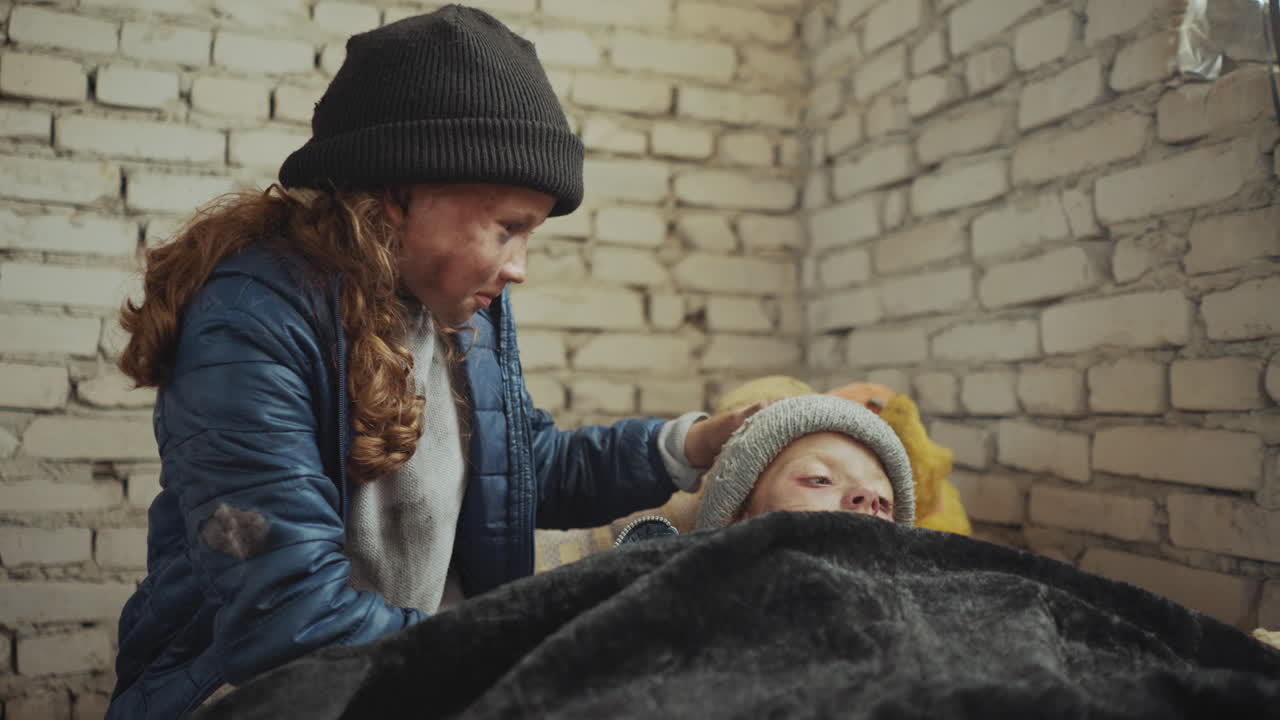 Tender moment as orphan girl with dirty face in blue jacket and black beanie gently touches forehead of sick sister resting under blanket in cold brick room, showing care, and emotional connection
