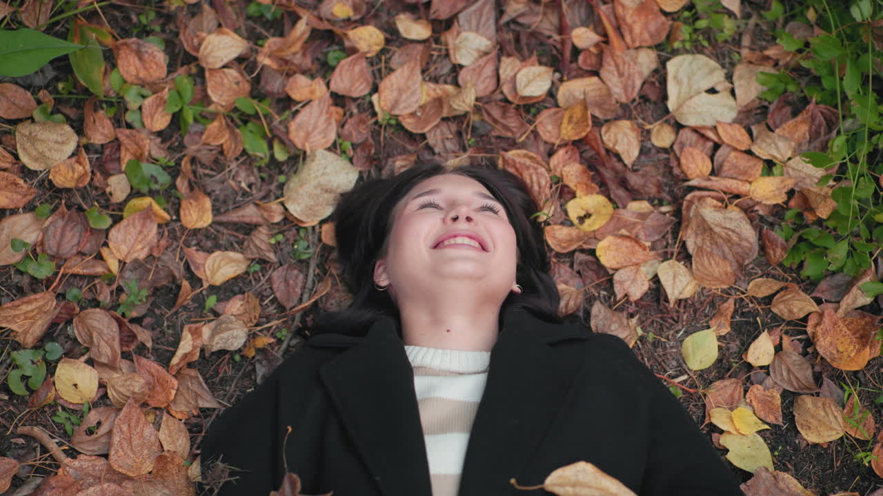 Smiling girl lies on forest floor among dry leaves, face up, smiling softly as single leaf drifts onto cheek, calm autumn mood, cozy coat, close view, tranquil season vibe, nature relaxation concept