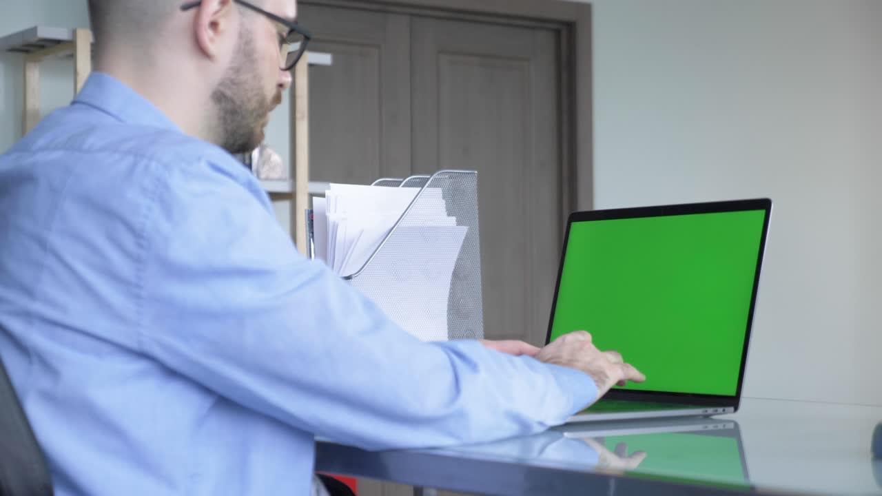 sobre el hombro de las manos del hombre escribiendo en una computadora portátil con pantalla de maqueta verde. en el fondo acogedora sala de estar con fuera de enfoque hombre