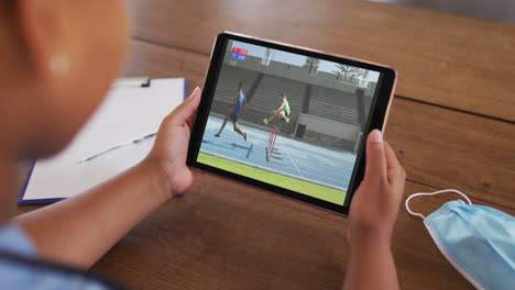 compuesto de una mujer sentada en una mesa con una máscara facial, viendo un evento de atletismo en la tableta