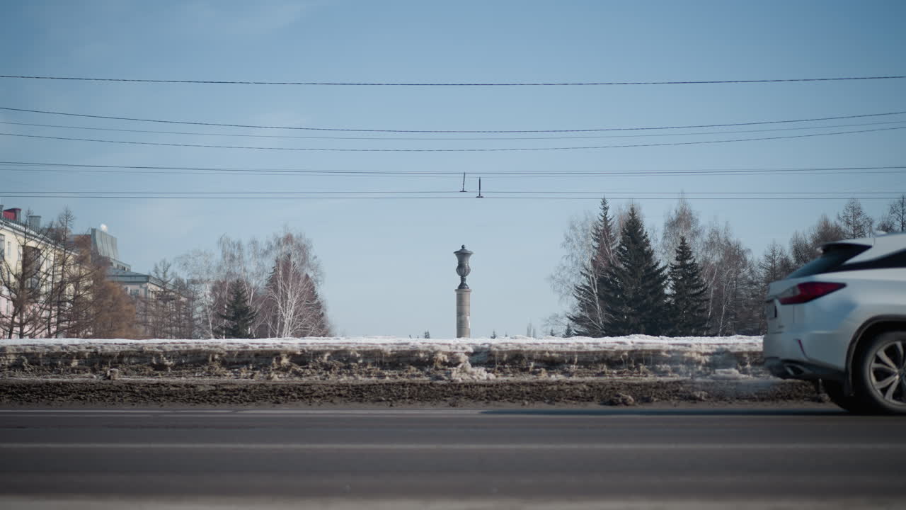 lower view of busy city road with cars moving on sides under wires, snow piled along median, trees flanking lanes, long pedestrian shadow crosses foreground, winter sky frames urban commute