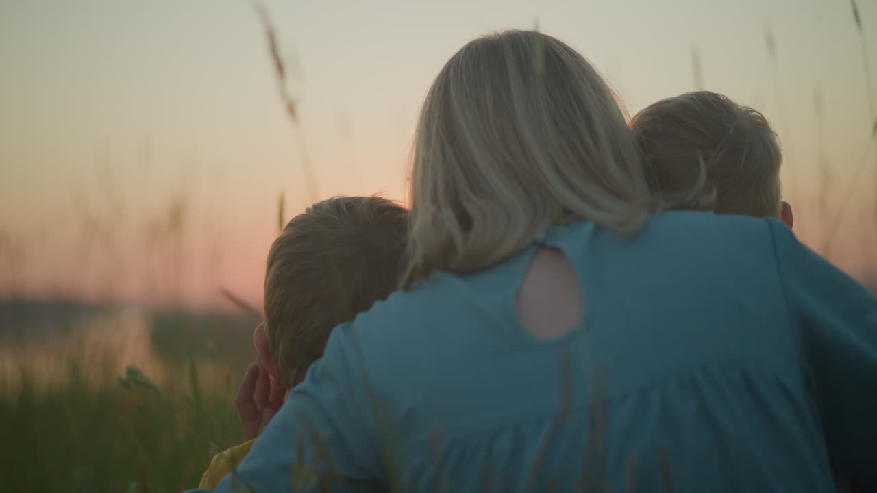 푸른 옷을 입은 어머니가 에 두 어린 아이와 함께 들판에 앉아 있으며, 그들은 모두 진정된 일몰을 지켜보고 있으며, 따뜻한 빛은 무대에 평화로운 빛을 고 있습니다.