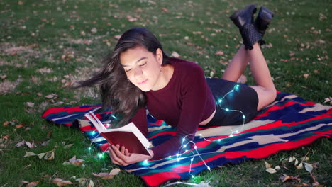 an attractive young woman reading a story book or novel in a park at twilight with lights glowing around her