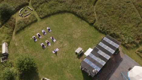 Drone-Shot-Of-Teacher-Leading-Group-Of-Mature-Men-And-Women-In-Class-At-Outdoor-Yoga-Retreat