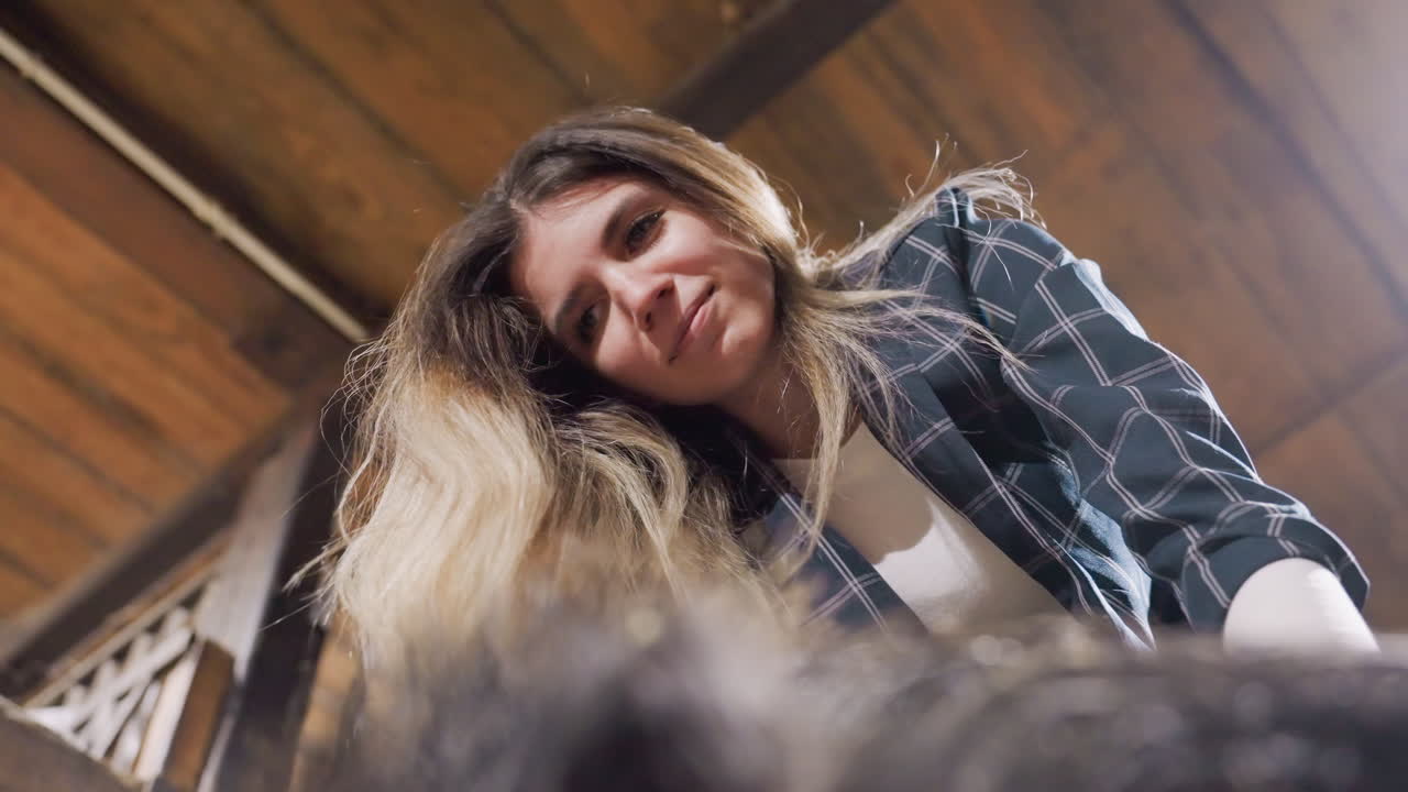 Mulher inclinada sobre a câmera mostrando inspeção de salão e estilo suave, retrato espontâneo em ângulo baixo com cabelo loiro, teto de madeira quente, mãos atentas da estilista, iluminação suave, cliente em ambiente íntimo