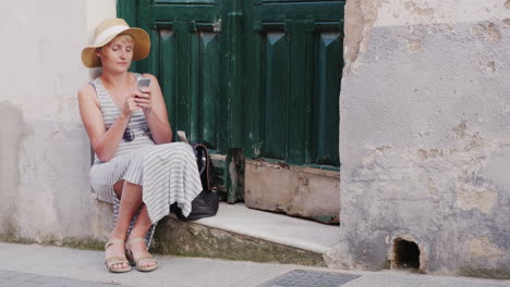 Woman-Tourist-Uses-Smartphone-He-Is-Sitting-On-The-Threshold-Of-The-Old-House-In-The-Resort-Town-Con