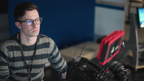 man in striped sweater working on camera in broadcast studio, adjusting settings on camera monitor, wearing glasses, focused on screen, professional video production environment