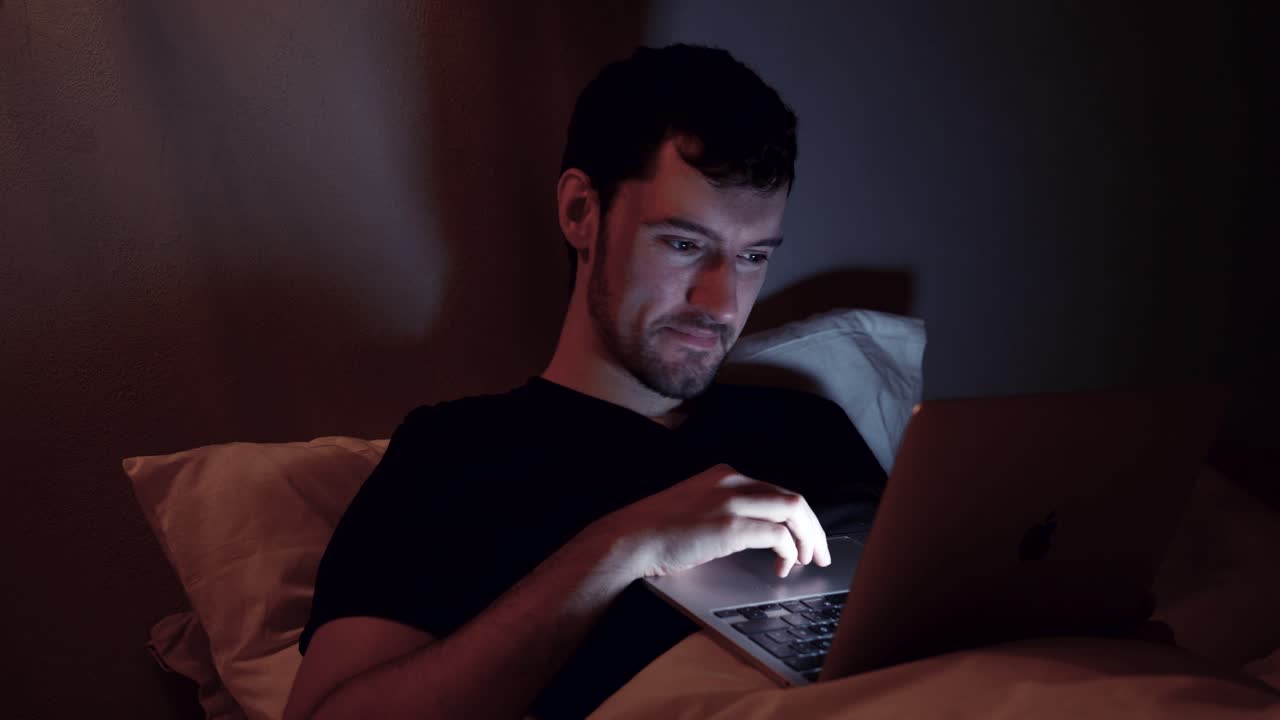 hombre con laptop en la cama riendo alegría emoción, noche noche internet en línea, trabajo tardío navegación web video viendo en el cuaderno antes de dormir
