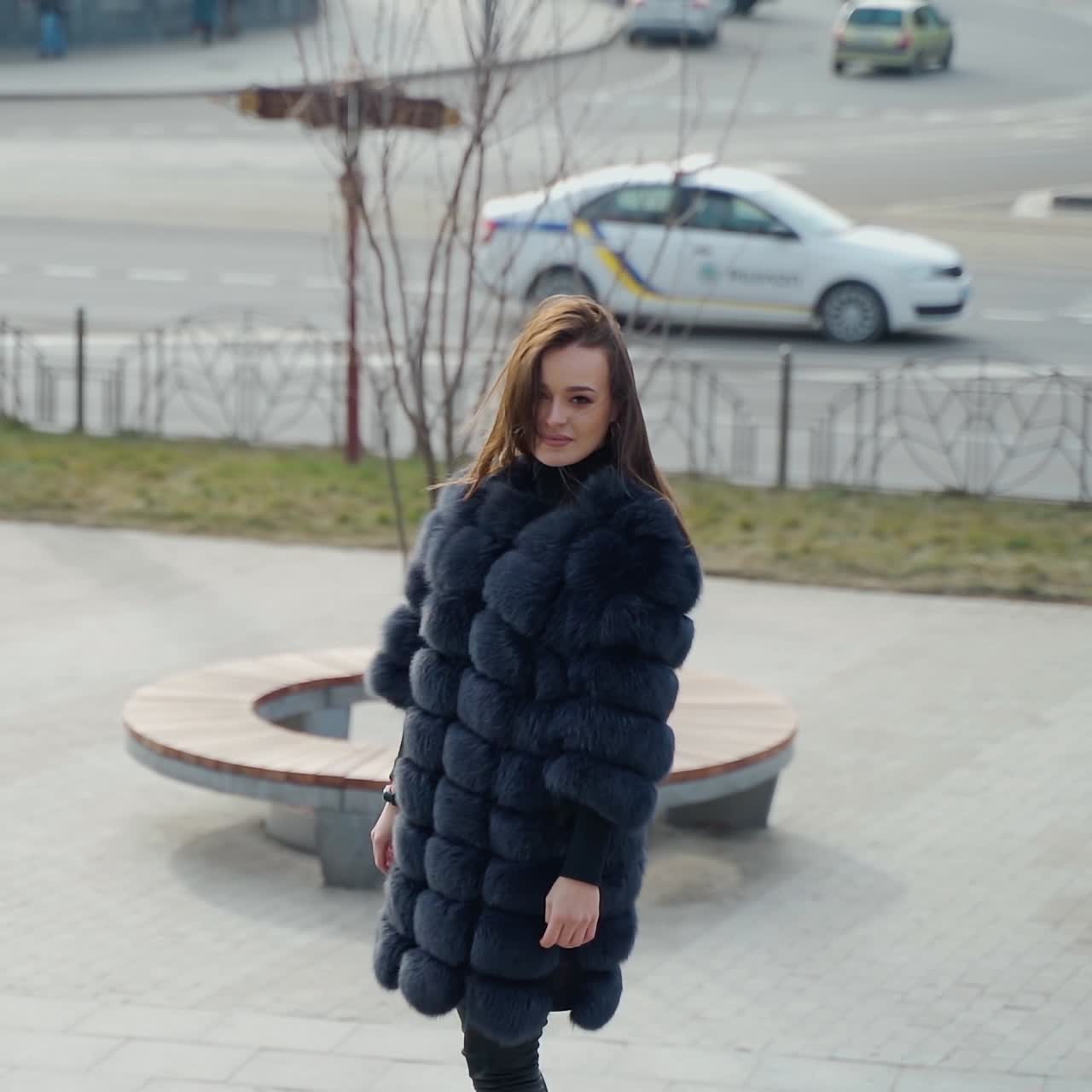 Girl in winter coat in the city. Beautiful woman in luxury fur coat posing on camera in the city park. Glamour model in the street. Slow motion.