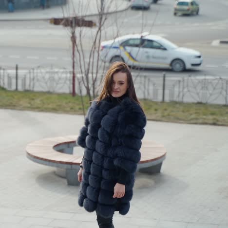 girl in winter coat in the city. beautiful woman in luxury fur coat posing on camera in the city park. glamour model in the street. slow motion.