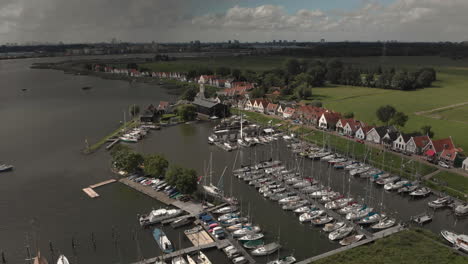 Panorámica-Aérea-Que-Muestra-La-Zona-Del-Puerto-Marítimo-Para-Embarcaciones-De-Recreo-Y-Veleros-Con-Casas-Típicas-Del-Pueblo-Holandés-De-Durgerdam-En-Durgerdammerdijk,-Cerca-De-Amsterdam,-Con-Nubes-Que-Proyectan-Una-Sombra-Profunda