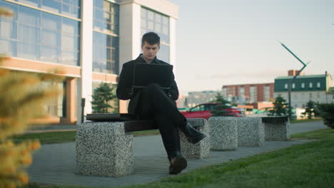 businessman seated on outdoor bench working on laptop with leg crossed in front of a modern office building. urban background includes greenery, vehicles, and clean architectural reflections