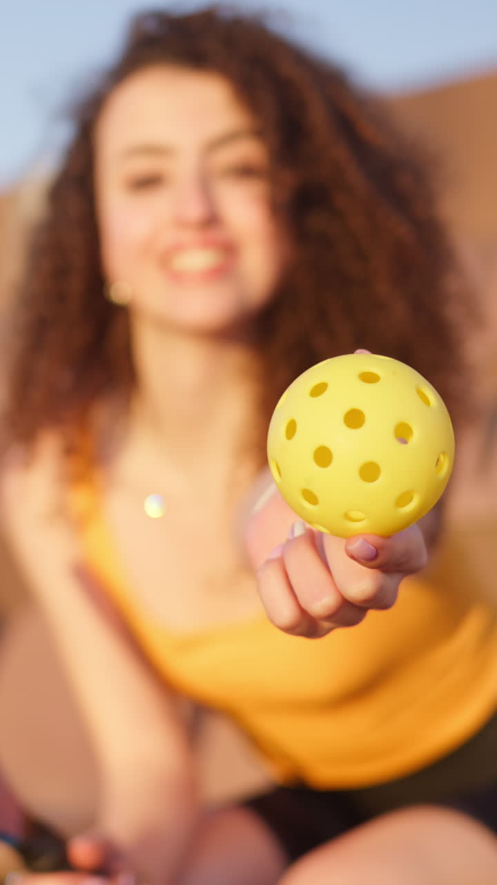 Close up of a yellow ball with a blurry view of a woman with curly hair sitting on a courtside seat, holding a pickleball racket. Vertical