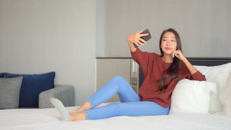 웃고 있는 아시아 여성은 집에 있는 침대에 앉아 스마트폰 카메라로 셀카를 찍는다