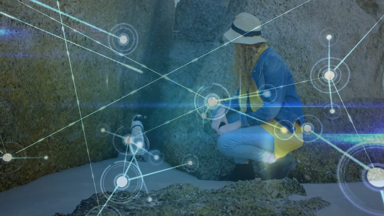 Donna accovacciata in una caletta rocciosa che sposta lo sguardo verso un pinguino, attivando una sovrapposizione di scansione tecnologica con nodi.
