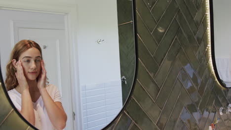 Happy-caucasian-woman-looking-in-mirror-and-touching-face-in-bathroom