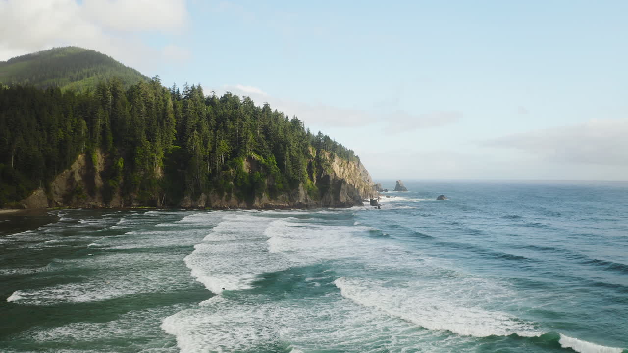 Sunlight hitting forested Oregon Coast hillside, aerial over Pacific Ocean waves