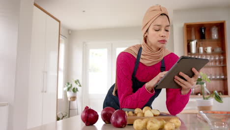 biracial woman in hijab using tablet preparing food in kitchen at home with copy space, slow motion