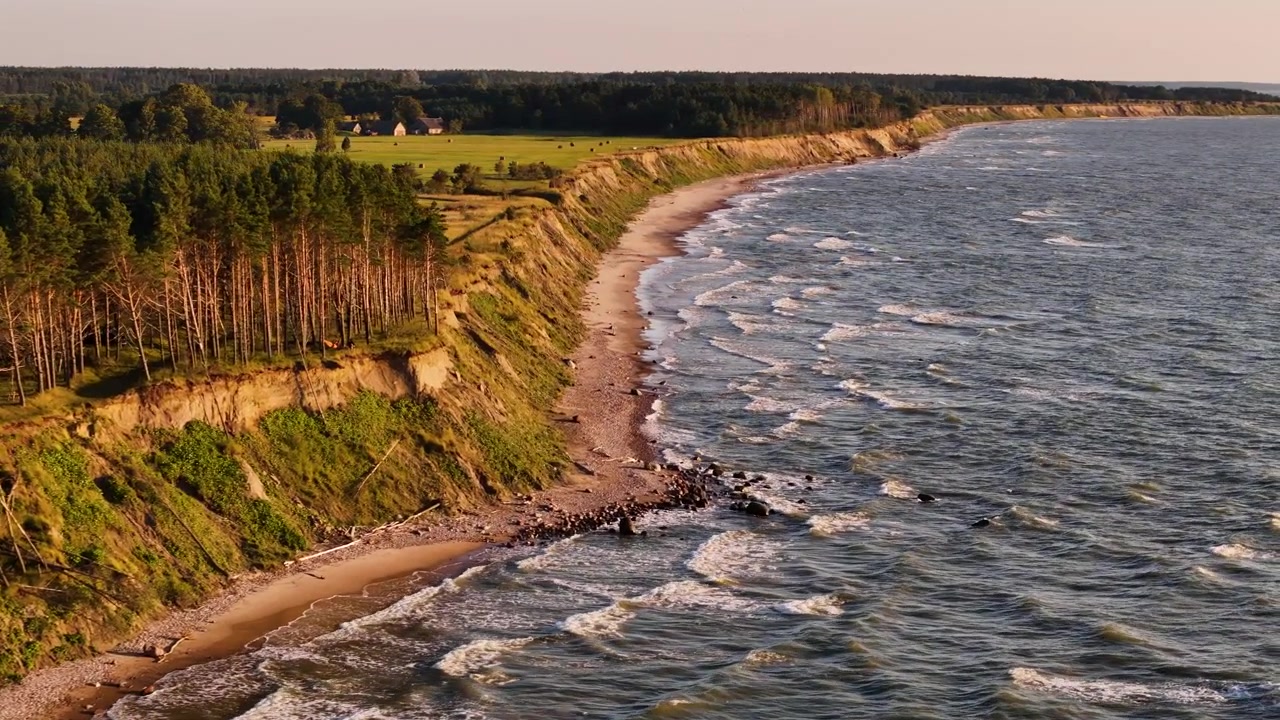Una vista aérea de un impresionante paisaje costero en Jurkalne Kurzeme. Un alto acantilado cubierto de vegetación verde domina una playa de arena y el vasto océano. Las olas rompen suavemente contra la orilla.