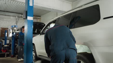 vista trasera de un ingeniero inspeccionando el neumático trasero de un jeep blanco usando la luz de una antorcha en una tienda de automóviles, con un colega en la parte delantera del vehículo, mostrando el mantenimiento del automóvil, el proceso de inspección de neumáticos
