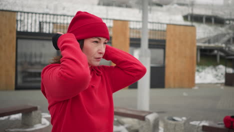 woman holding red beanie put it on during outdoor winter scene, flaunting hair with a relaxed expression and a slight smile, background features snowy urban landscape with modern structures