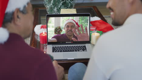 padre y hijo biraciales con sombreros de santa usando una computadora portátil para una videollamada de navidad con un hombre en la pantalla