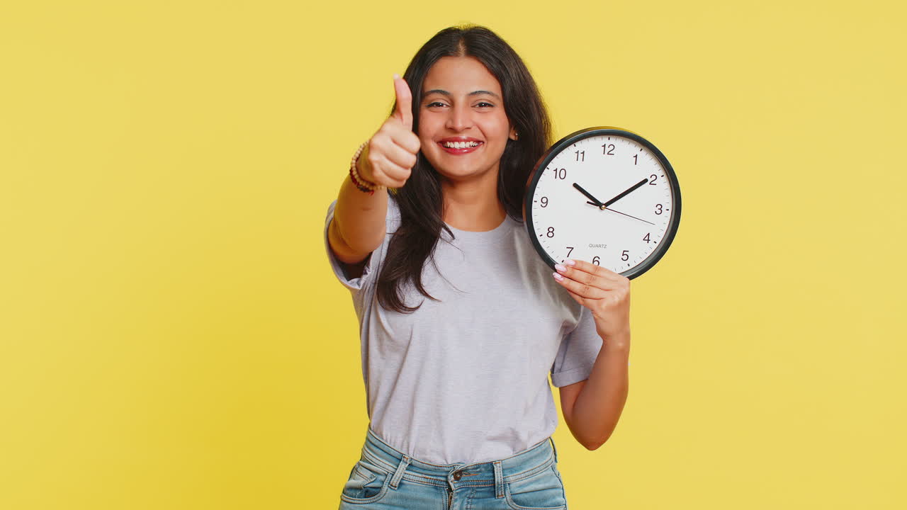 joven india mostrando la hora hora en el reloj de la oficina de la pared, ok, pulgar hacia arriba, aprobar, apresurarse, plazo