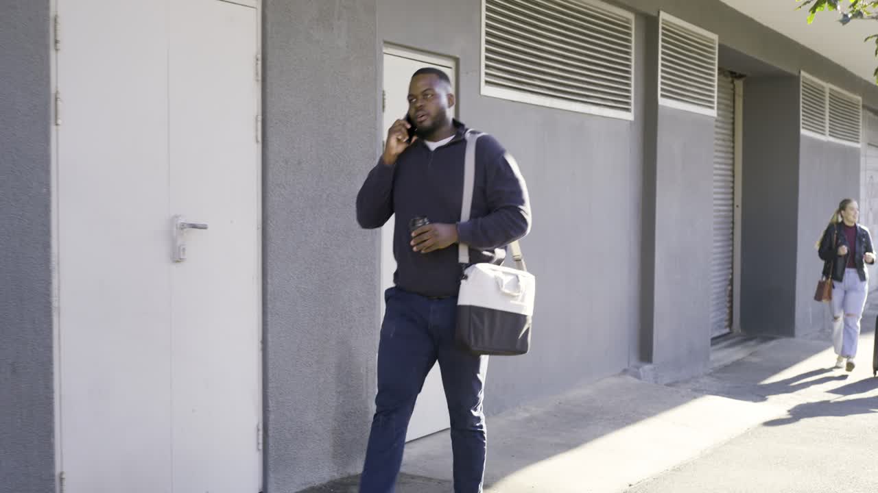 Vuxen man svarar i telefon, går på trottoaren, smuttar ur en kopp och håller en väska medan två andra drar resväskor.