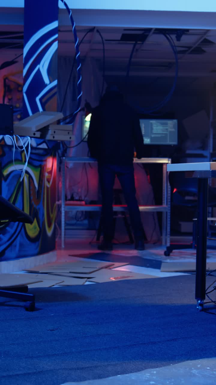 Video verticale di un hacker incappucciato che arriva in un rifugio sotterraneo nascosto illuminato al neon con graffiti